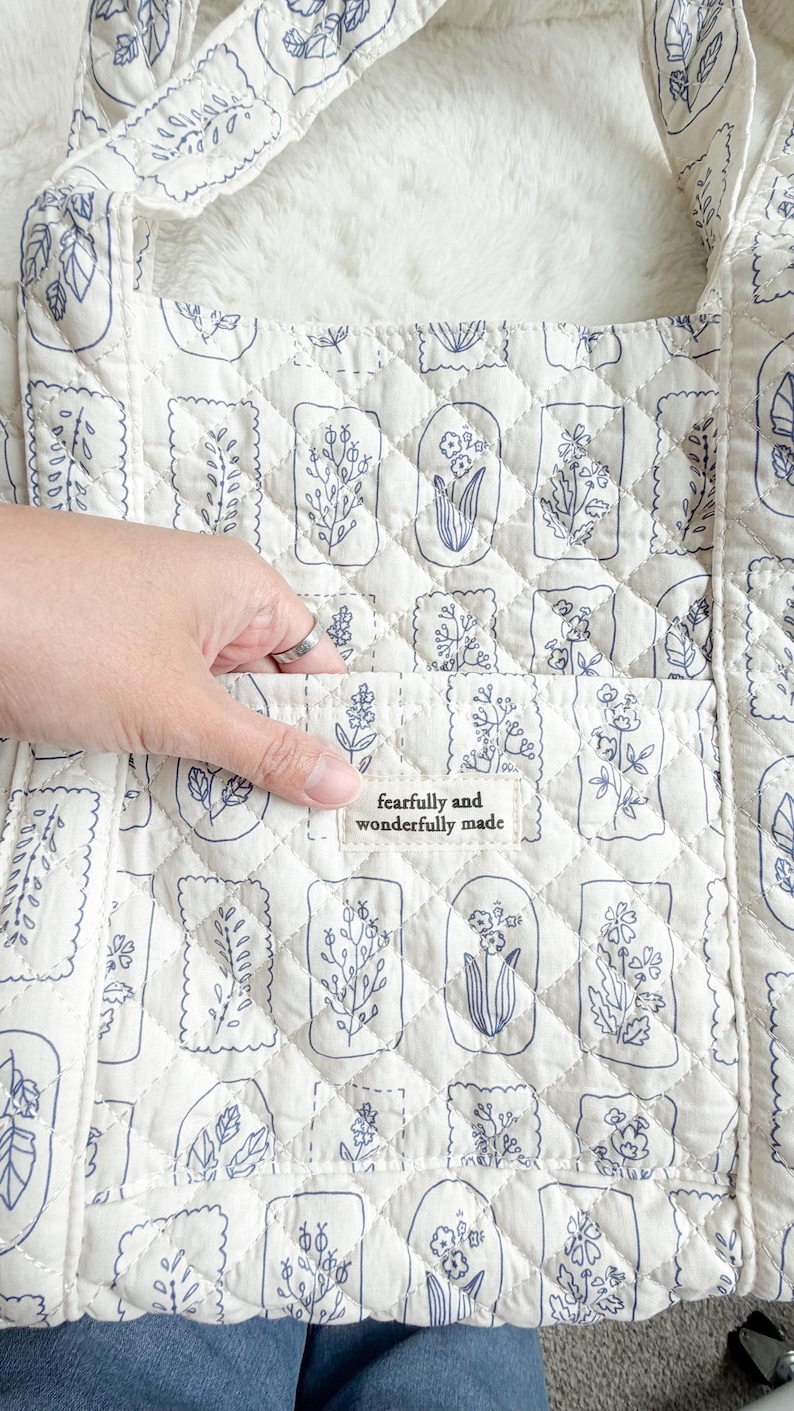 Puede incluir: Un bolso tote acolchado de color blanco roto con un patr&oacute;n repetitivo de l&iacute;neas azules de varias flores y hojas en marcos rectangulares. Una etiqueta de tela en el bolsillo frontal muestra el texto "fearfully and wonderfully made" en negro.