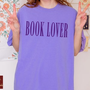 Op de afbeelding: Een paars T-shirt met de tekst "BOOK LOVER" in donkerpaars op de voorkant.