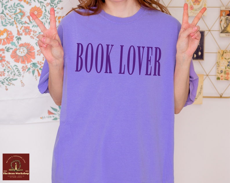 Op de afbeelding: Een paars T-shirt met de tekst "BOOK LOVER" in donkerpaars op de voorkant.