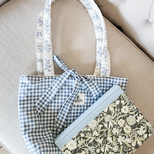 Puede incluir: Un bolso tote de cuadros vichy azul y blanco con cierre de lazo y asas acolchadas blancas con un estampado floral azul. Una etiqueta rectangular dice "BLUE BEAN WORKSHOP EST. 2021". Un libro con estampado floral verde y blanco descansa delante.