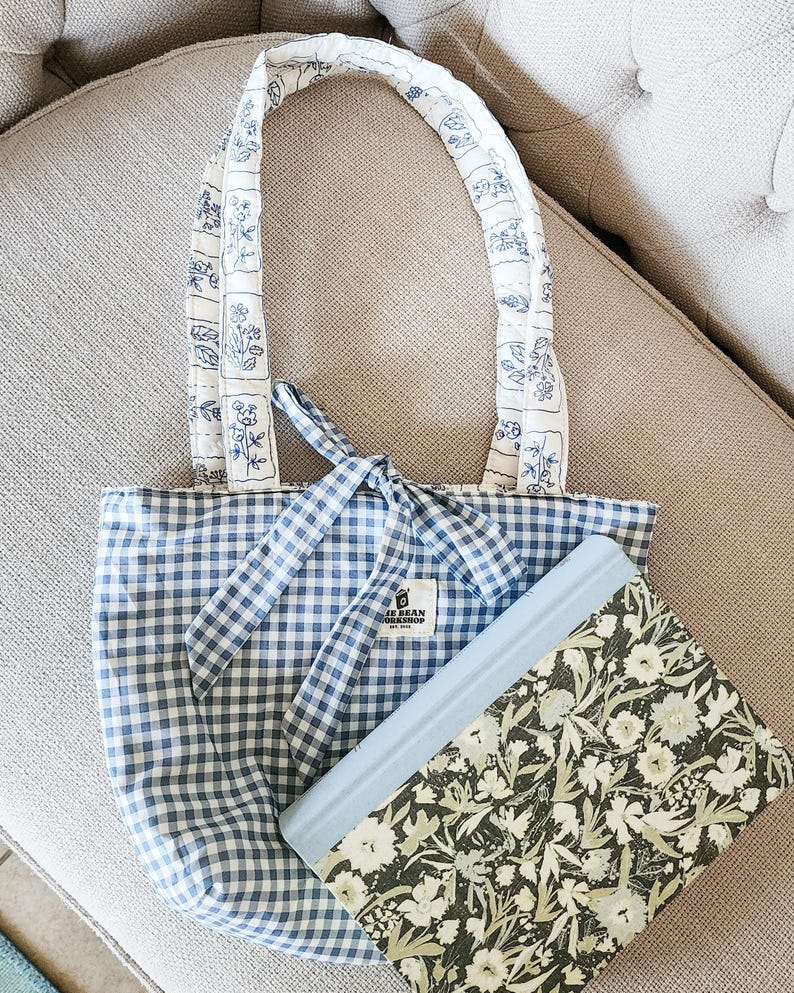 Puede incluir: Un bolso tote de cuadros vichy azul y blanco con cierre de lazo y asas acolchadas blancas con un estampado floral azul. Una etiqueta rectangular dice "BLUE BEAN WORKSHOP EST. 2021". Un libro con estampado floral verde y blanco descansa delante.