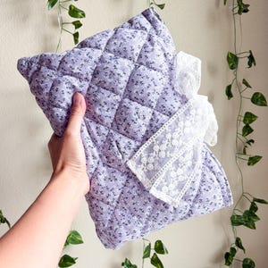 May include: A rectangular quilted pouch in a light lavender fabric featuring a small white floral pattern. The pouch is accented with a decorative white lace ribbon tied in a bow on the front, adding a textured detail to the soft, stitched surface.