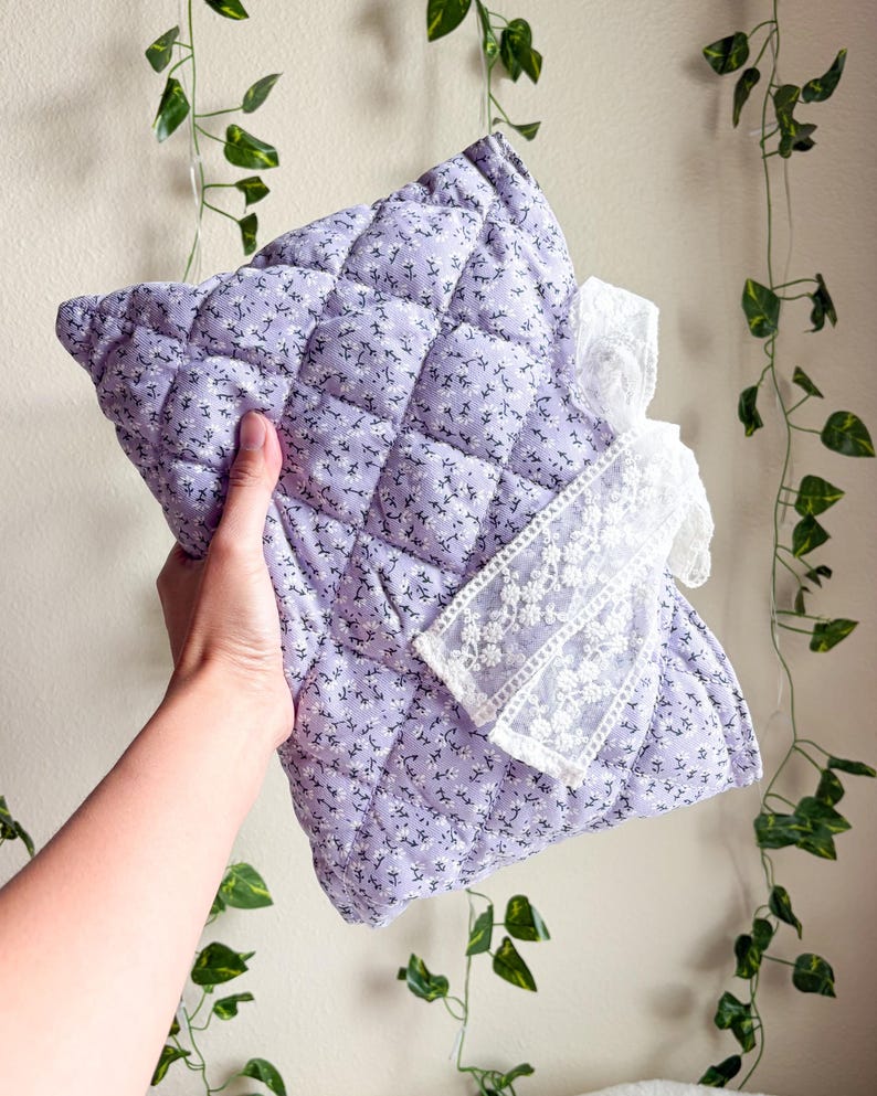May include: A rectangular quilted pouch in a light lavender fabric featuring a small white floral pattern. The pouch is accented with a decorative white lace ribbon tied in a bow on the front, adding a textured detail to the soft, stitched surface.