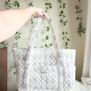 Puede incluir: Un bolso tote acolchado de tela color blanco roto con un patr&oacute;n floral repetitivo de l&iacute;neas azules. El bolso tiene dos correas para el hombro, bolsillos laterales y un bolsillo frontal central con una etiqueta que dice fearfully and wonderfully made.