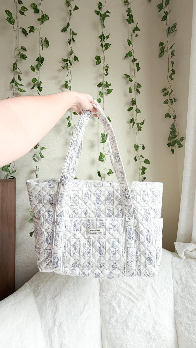 Puede incluir: Un bolso tote acolchado de tela color blanco roto con un patr&oacute;n floral repetitivo de l&iacute;neas azules. El bolso tiene dos correas para el hombro, bolsillos laterales y un bolsillo frontal central con una etiqueta que dice fearfully and wonderfully made.