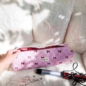 May include: Pink corduroy cosmetic bag with a bow pattern and the text "BOOKISH THINGS". A silver curling iron, a silver bow-shaped hair clip, and a curling iron are also visible.