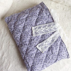 May include: A quilted fabric cover in a light purple hue featuring a small white floral pattern. The rectangular item is accented with a white lace ribbon tied in a bow on the right side, adding a decorative detail to the soft, padded textile surface.