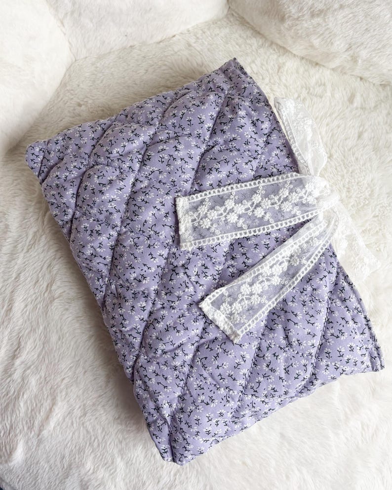 May include: A quilted fabric cover in a light purple hue featuring a small white floral pattern. The rectangular item is accented with a white lace ribbon tied in a bow on the right side, adding a decorative detail to the soft, padded textile surface.