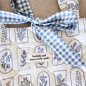 Puede incluir: Un bolso tote acolchado de tela blanca con un estampado bot&aacute;nico de l&iacute;neas azules. El bolso incluye una cinta de cuadros vichy azul y blanca atada en la parte superior y una etiqueta rectangular con el texto "fearfully and wonderfully made".