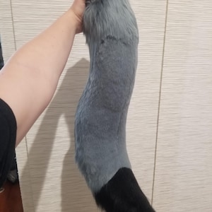 May include: A plush, gray and black faux fur tail. The tail is held up, showing a curved shape. The top portion is gray, transitioning to a black tip. The fur appears soft and fluffy, suitable for cosplay or costume use.