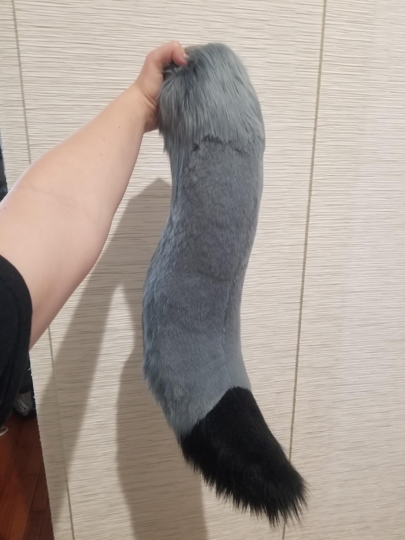 May include: A plush, gray and black faux fur tail. The tail is held up, showing a curved shape. The top portion is gray, transitioning to a black tip. The fur appears soft and fluffy, suitable for cosplay or costume use.