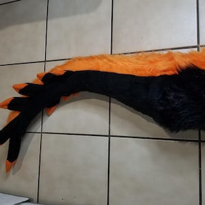 May include: A black and orange furry tail prop, likely for a costume. The tail is primarily black with an orange stripe along the top. The tip of the tail has three black and orange claw-like appendages.