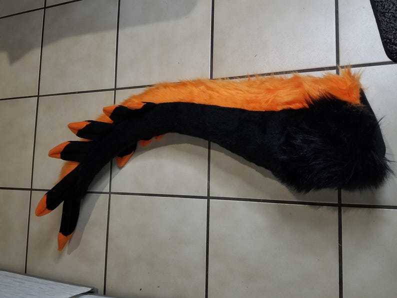 May include: A black and orange furry tail prop, likely for a costume. The tail is primarily black with an orange stripe along the top. The tip of the tail has three black and orange claw-like appendages.