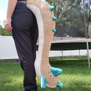 May include: A plush, beige and brown dragon tail with turquoise accents. The tail features a series of turquoise crystal-like shapes along the spine and at the tip. The tail is being held by a person wearing black pants and a navy blue shirt.