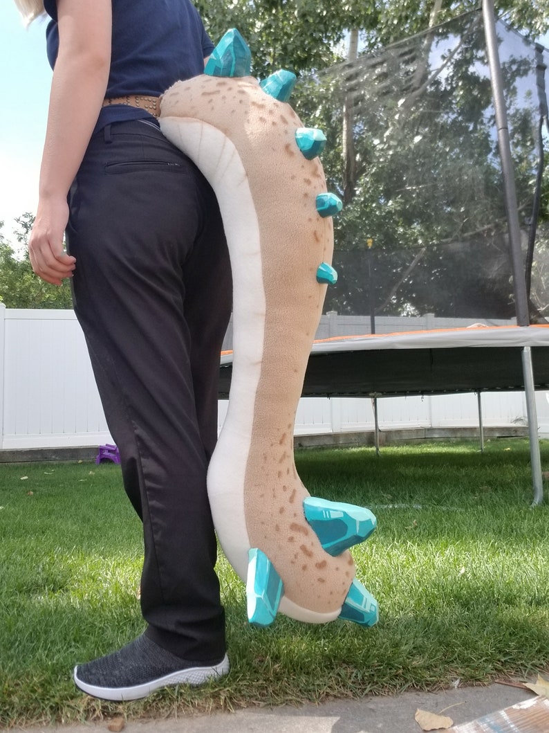May include: A plush, beige and brown dragon tail with turquoise accents. The tail features a series of turquoise crystal-like shapes along the spine and at the tip. The tail is being held by a person wearing black pants and a navy blue shirt.