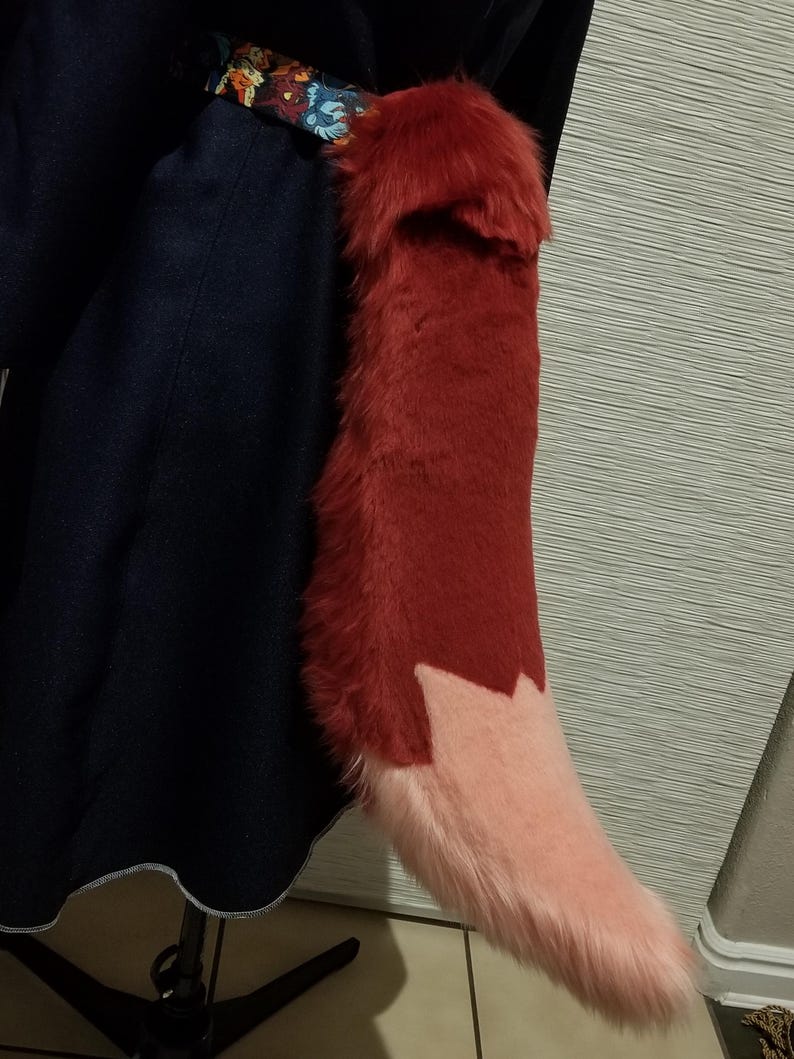 May include: A plush, red fox tail with a light pink tip. The tail is attached to a dark blue garment with a decorative floral patterned belt. The tail is made of soft, faux fur.