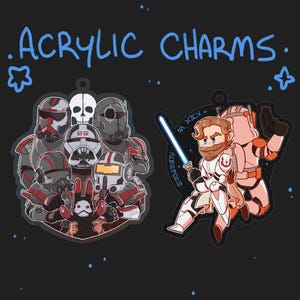 Pode incluir: Dois pingentes de acrílico com personagens do universo Star Wars. O primeiro pingente é um grupo de cinco soldados clone da 501ª Legião, com um esquema de cores preto e vermelho. O segundo pingente é uma representação de desenho animado do Mestre Jedi Obi-Wan Kenobi, segurando um sabre de luz azul, com um esquema de cores branco e laranja. O texto "501st" e "Kenobi" está escrito nos pingentes.