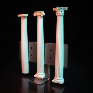 Op de afbeelding: Drie witte miniatuurkolommen met gegroefde details, gemonteerd op een metalen basis. De kolommen zijn in verschillende stijlen, een met een eenvoudige bovenkant, een met een decoratieve bovenkant en een met een meer versierde bovenkant.