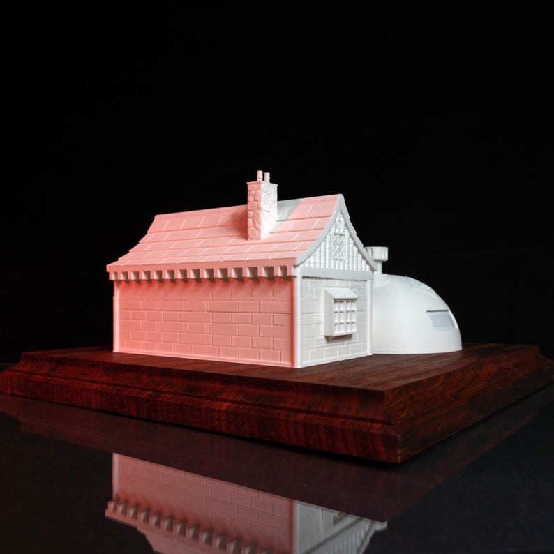 May include: White 3D printed model of a small house with a domed roof on a wooden base. The house has a chimney and a window.