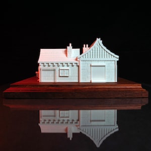May include: A white 3D printed model of a small house with a gabled roof and a chimney. The house is sitting on a wooden base.