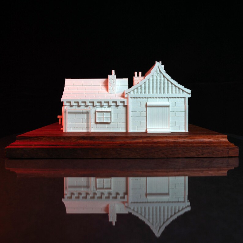 May include: A white 3D printed model of a small house with a gabled roof and a chimney. The house is sitting on a wooden base.