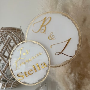 Könnte beinhalten: Zwei weiße runde Acrylschilder mit goldener Beschriftung. Das größere Schild sagt "B & L" und das kleinere Schild sagt "Zur Kommunion Stella".