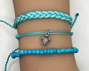 May include: A set of three turquoise bracelets with a silver turtle charm. The top bracelet is braided with white stripes, the middle bracelet is a simple string, and the bottom bracelet is made of small turquoise beads.