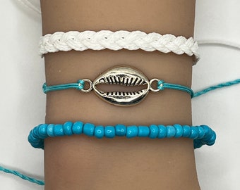 May include: Set of four bracelets featuring a white braided bracelet, a turquoise string bracelet with a silver seashell charm, and two turquoise beaded bracelets.