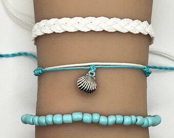May include: Three bracelets stacked on a wrist. The top bracelet is white and braided. The middle bracelet is white and blue with a silver seashell charm. The bottom bracelet is turquoise blue and made of small beads.