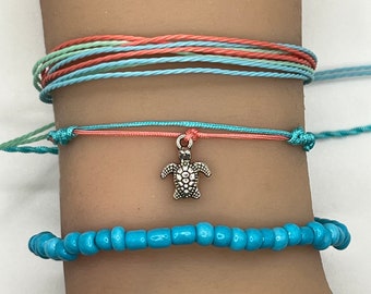 May include: A set of three bracelets featuring a turquoise beaded bracelet, a turquoise and coral string bracelet with a silver turtle charm, and a multi-colored string bracelet.