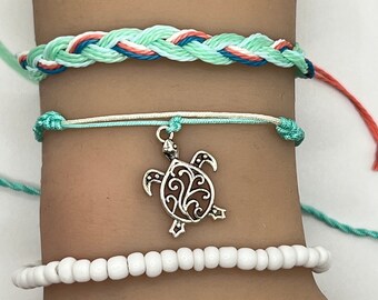 May include: A set of three bracelets featuring a braided turquoise and white string bracelet, a turquoise string bracelet with a silver turtle charm, and a white bead bracelet.