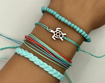 May include: A set of four turquoise and blue bracelets with a silver turtle charm. The bracelets are made of different materials, including braided cord and beads.