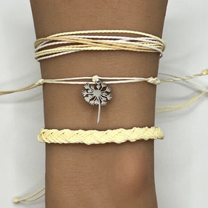 May include: A set of three yellow and white string bracelets with a silver dandelion charm. The bracelets are layered on a person's wrist.
