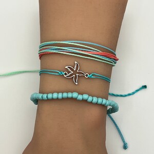 May include: A set of three bracelets featuring a turquoise blue beaded bracelet, a turquoise blue string bracelet with a silver starfish charm, and a multi-colored string bracelet with turquoise blue, light green, and coral pink strings.