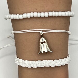 May include: Three white bracelets with a ghost charm. The top bracelet is made of white beads, the middle bracelet is a white string bracelet, and the bottom bracelet is a white braided rope bracelet.