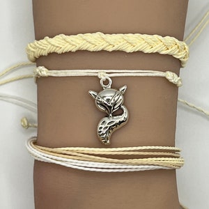 May include: A set of four bracelets with a silver fox charm. The bracelets are made of braided rope in shades of white and cream.