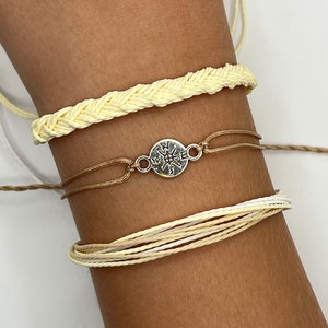 May include: A set of three bracelets featuring a braided yellow cord, a brown cord with a silver compass charm, and a multi-layered white and brown cord.