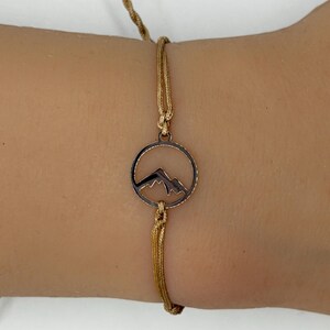 May include: A gold-colored bracelet featuring a circular pendant with a mountain range silhouette. The bracelet is made of a woven cord and is secured with adjustable knots. The mountain pendant is a dark bronze color.