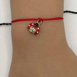 May include: Red string bracelet featuring a gold-colored ladybug charm with red enamel wings, black accents, and a clear crystal center. The bracelet is tied with a simple knot.