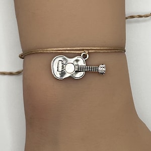 May include: A tan string bracelet with a silver-colored guitar charm. The guitar charm has detailed features, including the sound hole and strings. The bracelet is tied with a simple knot.