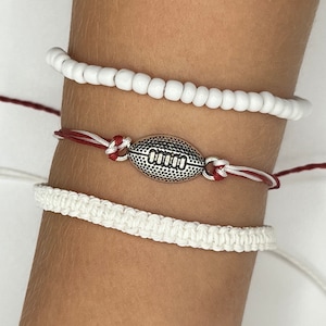 May include: Three white bracelets on a wrist. The top bracelet is a string of white beads. The middle bracelet is a red and white string with a silver football charm. The bottom bracelet is a white braided cord.