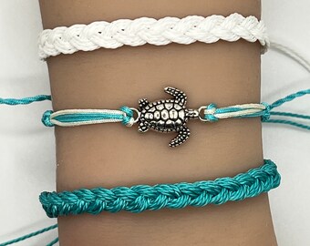 May include: Three braided friendship bracelets with a silver turtle charm. The top bracelet is white, the middle bracelet is white and light blue, and the bottom bracelet is teal.