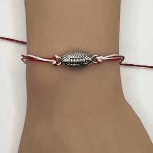 May include: A silver football charm bracelet with red and white string. The football charm is detailed with lines and stitching. The bracelet is tied on the wrist with the red and white string.