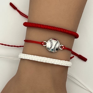 May include: Three bracelets on a wrist. The top bracelet is red, the middle bracelet is red with a silver baseball charm, and the bottom bracelet is white.