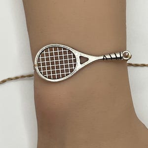 May include: A silver-toned tennis racket charm bracelet. The racket charm has a brown net design and is strung on a tan cord. The bracelet is worn on a wrist.