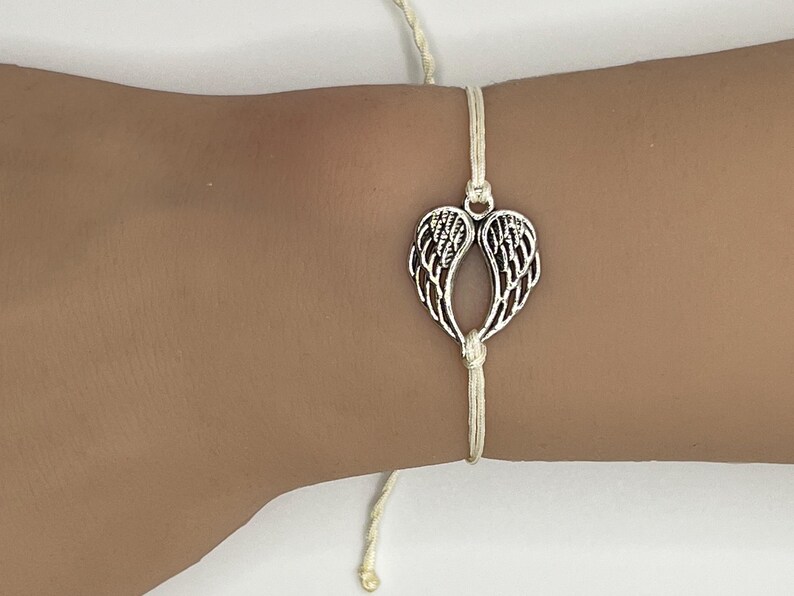 Puede incluir: Una pulsera con un dije de alas plateadas. El dije tiene forma de dos alas unidas en la parte superior e inferior, ensartadas en un cord&oacute;n color crema. La pulsera est&aacute; atada a la mu&ntilde;eca.