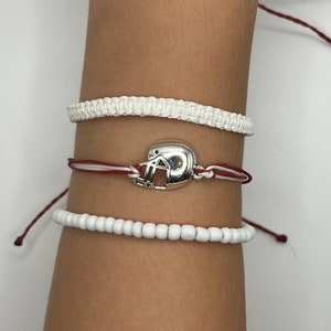 May include: Three bracelets on a wrist. The top bracelet is white and braided. The middle bracelet is red and white with a silver football helmet charm. The bottom bracelet is white and made of beads.