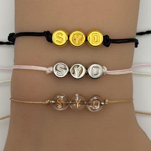 May include: Three personalized bracelets with the letters "SYD" are displayed. The top bracelet has gold beads on a black cord. The middle bracelet features silver beads on a pink cord. The bottom bracelet has clear beads on a tan cord.
