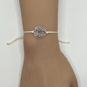 Puede incluir: Una pulsera con dije de alas de color plateado. El dije tiene forma de dos alas unidas en el centro. La pulsera est&aacute; hecha de un cord&oacute;n de color crema. La pulsera se lleva en la mu&ntilde;eca.