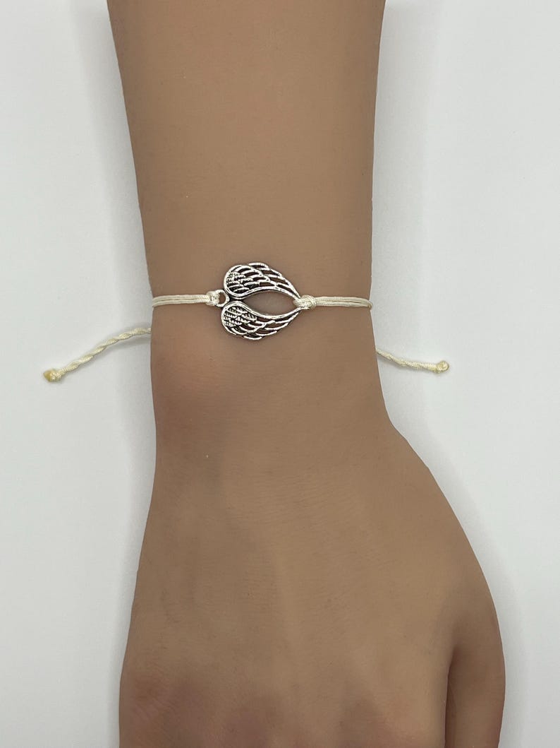Puede incluir: Una pulsera con dije de alas de color plateado. El dije tiene forma de dos alas unidas en el centro. La pulsera est&aacute; hecha de un cord&oacute;n de color crema. La pulsera se lleva en la mu&ntilde;eca.